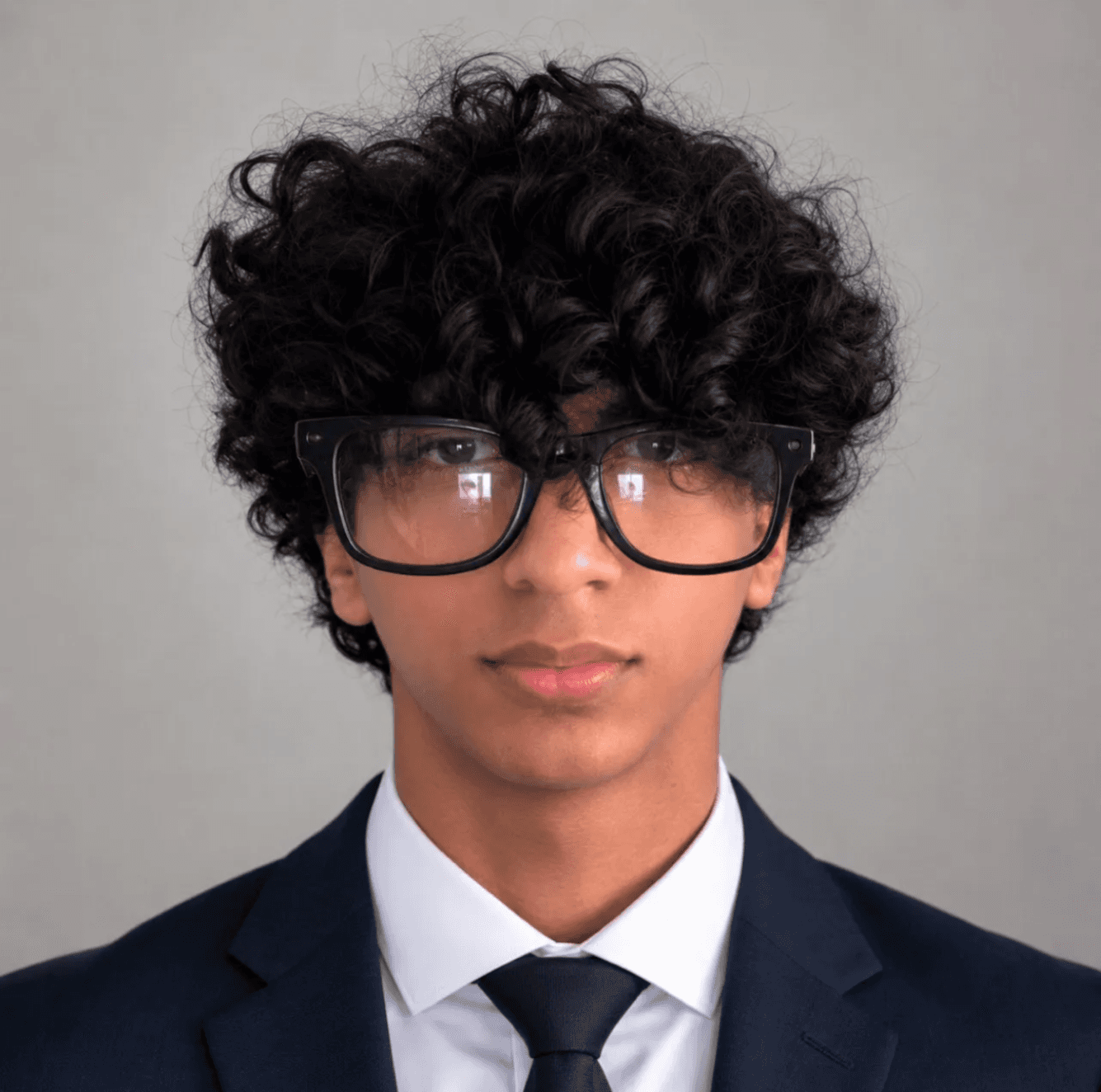 Portrait of Rishab Ali Muhammad in a navy suit, white shirt, and dark tie, wearing black-framed glasses